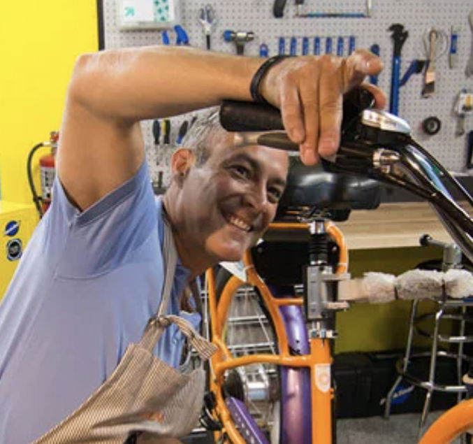 Pedego- A person wearing an apron smiles while fixing a bicycle in a workshop with tools on a pegboard in the background.
