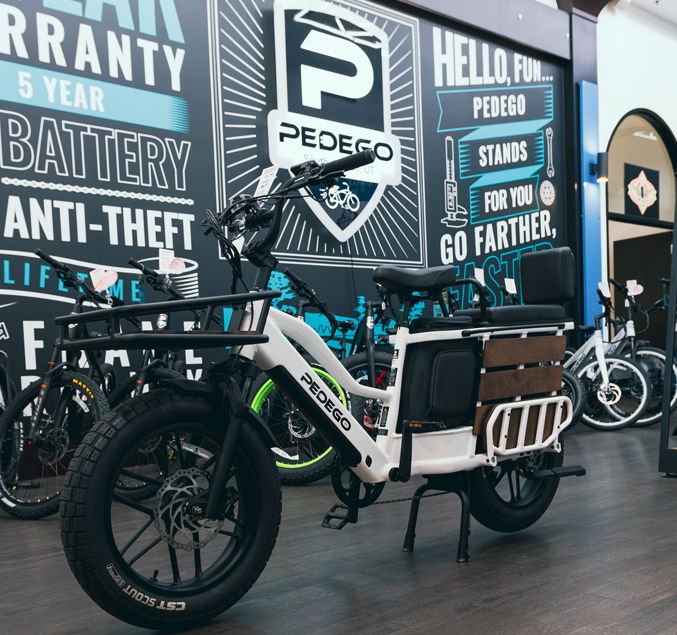 Pedego- A white electric bike is displayed indoors against a backdrop featuring the Pedego logo and promotional text including "5 Year Warranty," "Anti-Theft," and "Hello, Fun..." Other bikes are visible in the background.