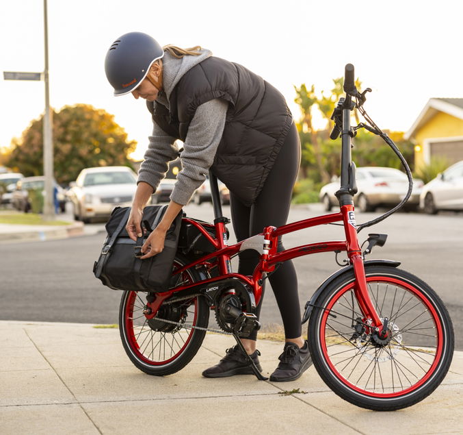 Pedego- A person wearing a helmet and a puffy vest secures a bag onto a red folding bicycle on a sidewalk.