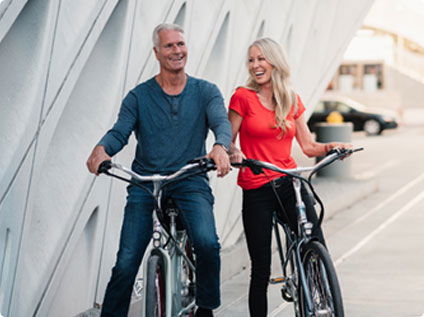 Pedego- A man and a woman are riding bicycles side by side on a city street, smiling and engaged in a lively conversation. The background features a modern architectural wall.