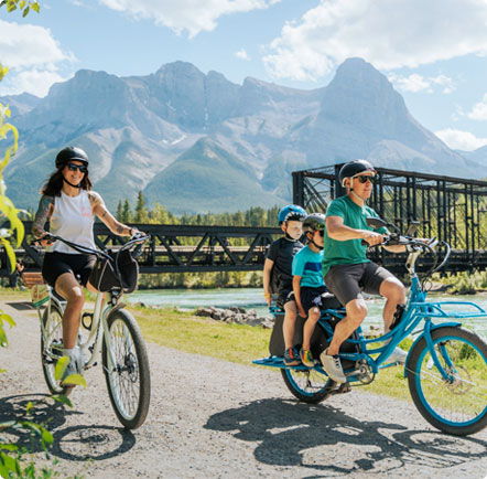 Pedego- A family rides bicycles on a gravel path near a scenic mountain range. An adult woman and a man with two children and a child seat can be seen riding.