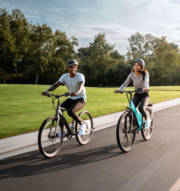 Pedego- Two people wearing helmets ride bicycles on a paved path through a park, with green trees and grass in the background.