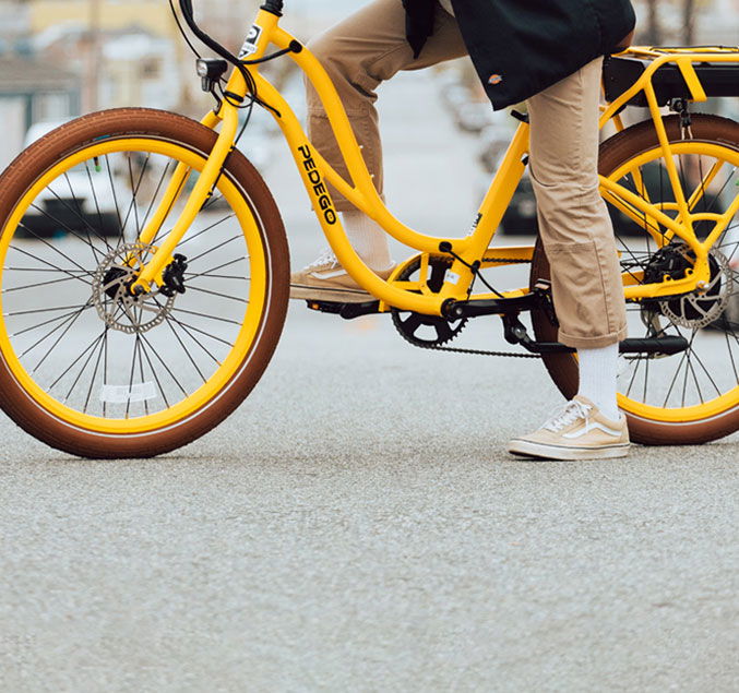 Pedego- Person standing next to a yellow Pedego bicycle, wearing beige pants, white socks, and tan sneakers, on a paved road.