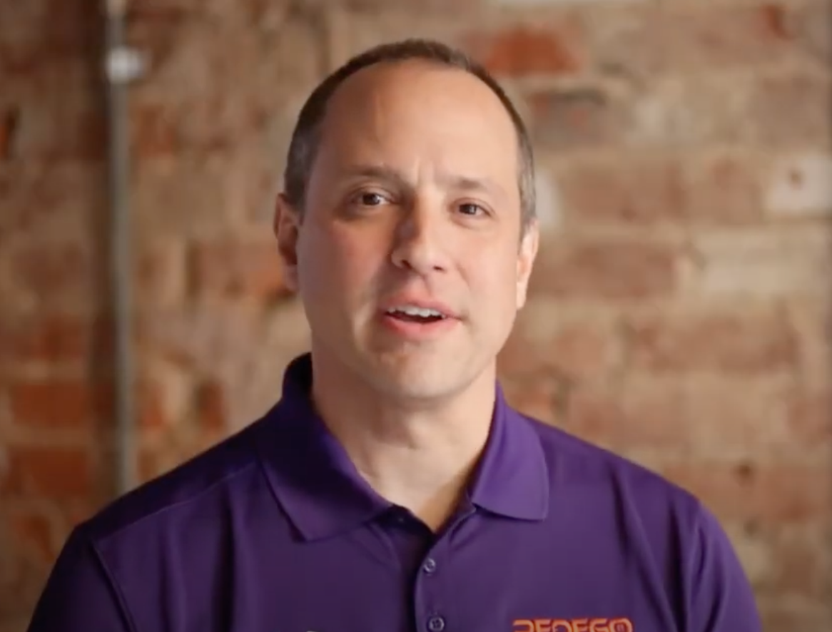 Pedego- A man wearing a purple polo shirt stands in front of a brick wall, looking at the camera.