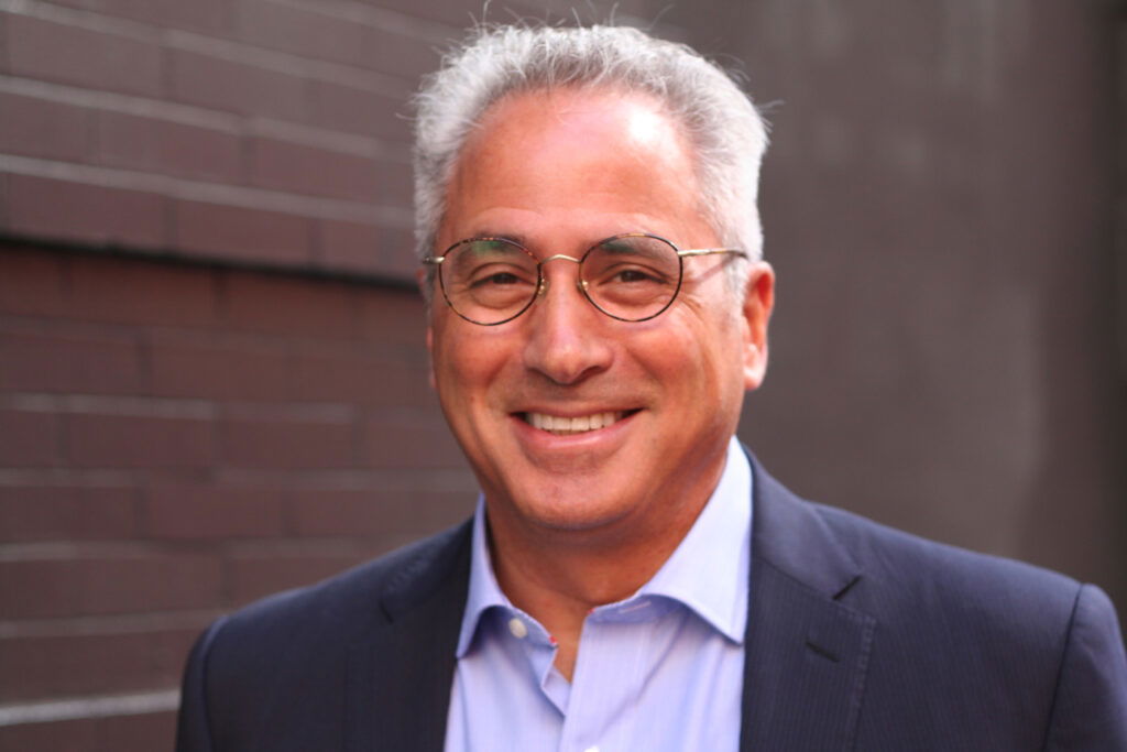 Pedego- Larry Pizzi - A man with short gray hair and glasses smiles while wearing a navy blazer and light blue shirt. He stands in front of a brick wall.